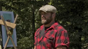 Een foto van boswachter Tim. Hij staat buiten en kijkt opzij. Hij heeft een rood-zwart geruit overhemd aan en een pet op. Op de achtergrond zie je bomen en een bord met de foto van een uil erop.