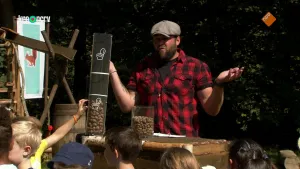 Een foto van boswachter Tim die buiten achter een tafel staat. Hij heeft een doorzichtige buis vast met daarin eikels. Hij is iets aan het vertellen. Op de voorgrond zie je kinderen.