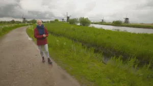 Een foto van presentator Janouk. Ze loopt op een pad, richting de camera. Naast het pad is grasland en daarnaast water. Op de achtergrond zie je vijf oude molens. 