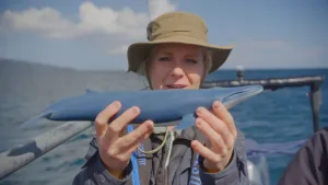 Een foto van presentator Janouk. Ze staat in een bootje en toont de kijker een schaalmodel van een Blauwe Vinvis. Ze kijkt daarbij in de camera.