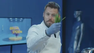 Een foto van hersenwetenschapper Job van den Hurk in de Tv-studio van Brainstorm. Hij houdt een bosje groene sprietjes vast en toont die aan de camera. Op de achtergrond zie je een pop van een mens, met een hoofd dat open is aan de bovenkant.