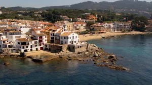 Een foto van een stadje aan de zee. Alle huizen zijn wit met oranje daken. Vooraan is een stukje rotsige grond dat half in het water ligt. Daarachter ligt een klein strand. Op de achtergrond zijn veel bomen en wat heuvels. 