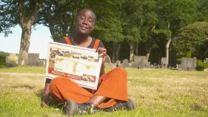 Een foto van presentator Tirsa. Ze zit buiten in het gras en houdt een plattegrond vast. Ze kijkt in de camera. Op de achtergrond zie je grafstenen en bomen.