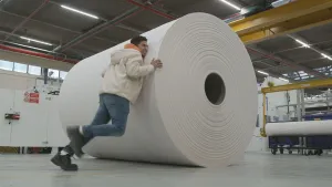 Een foto van presentator Nizar in een fabriekshal. Je ziet een enorm grote rol papier en Nizar duwt daar tegenaan. Het kost hem veel moeite zo te zien. 