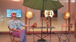 Een foto van presentator Janouk. Ze zit in de Tv-studio van Brainstorm op een stoel. Naast haar staan een tafel en stoel en een parasol. Janouk heeft een zonnebril op en kijkt schuin omhoog. Op de tafel staan drankjes. 