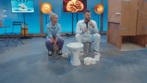 Een foto van Janouk en een mannelijke presentator in de Tv-studio van Brainstorm. Ze zitten allebei op een emmer. Voor hen staat een wc-pot en er liggen wc-rollen op de grond. Achter hen zie je een plaatje van hersenen op een beeldscherm.