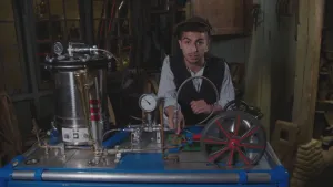 Een foto van presentator Nizar. Hij staat in een schuur, achter een stoommachine. Hij wijst naar een onderdeel van die machine en kijkt in de camera. Je ziet wielen en slangen en buizen op die stoommachine.