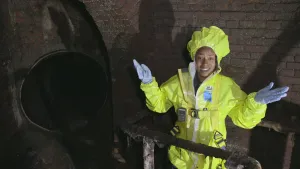 Een foto van presentator Eva. Ze heeft een geelgekleurd beschermend pak aan. Ze staat namelijk in een riool. Het ziet er vies uit. Links in beeld zie je een groot donker gat.