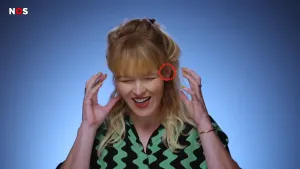 Een foto van de presentator op een kale achtergrond. Ze houdt haar handen omhoog richting haar oren, alsof ze pijn aan haar oren heeft. 