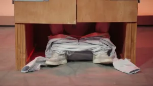 Een foto van de onderkant van een houten WC-hokje. Onder de deurtjes door is de WC-pot te zien, en de benen van iemand die op de WC zit. Zijn overall is naar beneden geschoven rond zijn enkels.