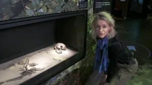 Een foto van presentator Janouk. Ze staat in een museum. Naast haar zie je het skelet van een mens. Men noemt haar Trijntje. Janouk praat in de camera. 