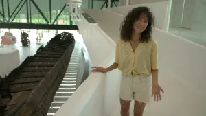 Een foto van presentator Serah. Ze staat in een museum en praat in de camera. Naast haar zie je een heel oud Romeins schip dat opgegraven is. 