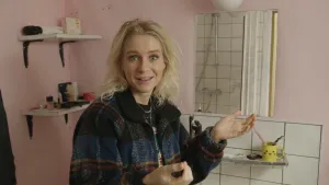 Een foto van presentator Janouk. Ze staat in een badkamer en praat in de camera. De muren zijn zachtroze van kleur. Het is een badkamer die zelf in elkaar geknutseld is.