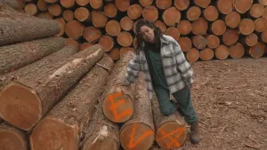 Een foto van presentator Eva. Ze is omringd door tientallen afgezaagde boomstammen. Ze leunt daar tegenaan. Op drie van boomstammen zijn de letters E V A geschreven. 