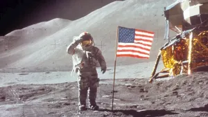 Een foto van de maanlanding. Een persoon in een ruimtepak staat naast een Amerikaanse vlag in een kaal landschap. Op de achtergrond staat een ruimteschip en daarachter zijn bergen. 