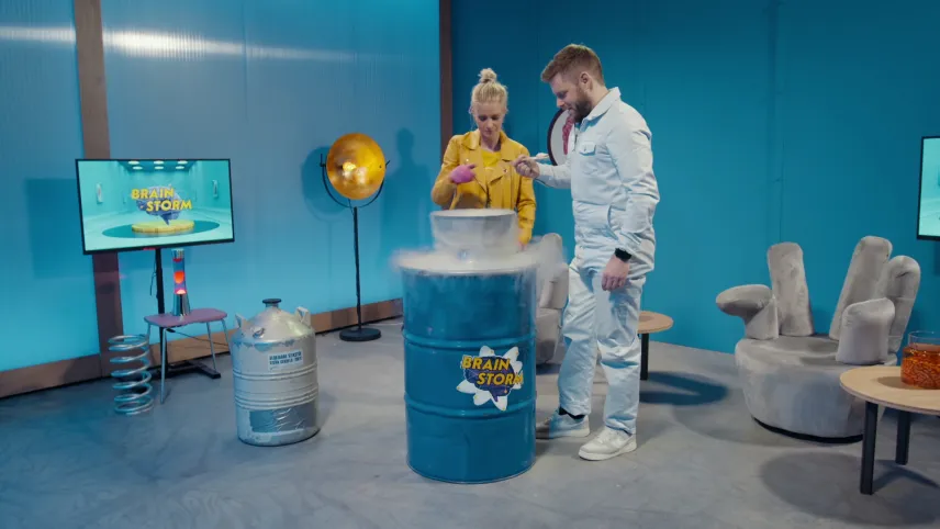Een foto van Janouk en prof die bij een blauw stalen vat met de tekst "Brainstorm" erop staan. Er staat een kom op met ijs erin waar koude lucht vanaf komt. Janouk heeft er ijskoude vloeibare stikstof in gedaan. Ze hebben een lepel met ijs erop vast.