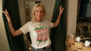 Een foto van Janouk die uit een paskamer komt. Ze heeft de twee gordijnen die ze open heeft gedaan vast. Ze draagt een wit t-shirt met het logo van Het Klokhuis. Achter haar hangt een spiegel in de paskamer en rechts achter een balie hangt kleding.
