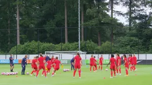 Een foto van het Nederlandse vrouwenvoetbalelftal dat aan het trainen is op een voetbalveld. De oranjevrouwen dragen geen oranje, maar rode t-shirts, broek en sokken en zijn de bal in groepjes naar elkaar aan het overspelen. Vier trainers kijken toe.