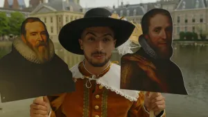 Een foto van presentator Nizar bij de Hofvijver in Den Haag. Hij heeft zestiende-eeuwse kleding aan. Hij houdt twee foto's op een bordje vast. De linkerfoto toont Johan van Oldenbarnevelt. De rechterfoto toont Prins Maurits van Oranje.