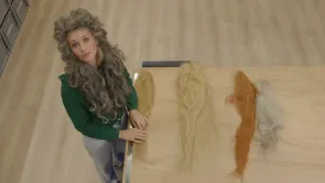 Een foto van presentator Janouk. Ze heeft een grote pruik op en kijkt in de camera. Voor haar liggen vier pruiken op tafel. Die pruiken hebben allemaal verschillende kleuren.