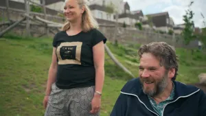 Een foto van een vrouw en een man. Ze zijn buiten en kijken in dezelfde richting. Ze glimlachen allebei. Op de achtergrond zie je een heuvel met daarop huizen. 