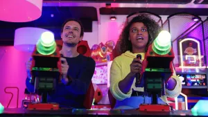 Een foto van presentator Quinty. Ze zit in een game hal en heeft soort elektronisch geweer in haar handen. Naast haar zit een man die ook zo'n apparaat vasthoudt. Ze kijken allebei aandachtig vooruit. 