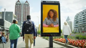 Een foto van een reclamezuil in een stad. Op die zuil is een foto van Quinty te zien. Naast die zuil loopt Quinty ook, samen met een man. Je ziet hen op de rug. Ze kijkt naar die reclamezuil.