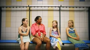 Een foto van presentator Quinty. Ze zit in de kleedruimte van een zwembad, samen met drie kinderen. De kinderen hebben zwemkleren aan en een handdoek om. Quinty praat met hen.
