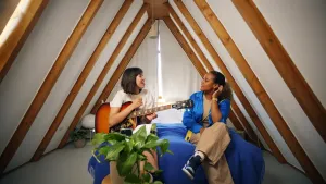 Een foto van zanger Meau en presentator Quinty. Ze zitten in een zolderkamer met een puntdak en praten met elkaar. Ze zitten op een bed. Meau heeft een gitaar op schoot. 