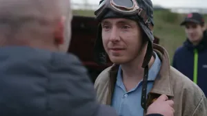 Een foto van presentator Pieter. Hij heeft een ouderwetse leren race-muts met bril op zijn hoofd. Hij is in gesprek met een man.