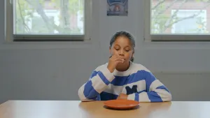 Een foto van een meisje van ongeveer tien jaar oud. Ze zit aan een tafel en stopt een marshmallow in haar mond. Ze kijkt naar een bord dat op tafel ligt.