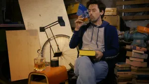 Een foto van presentator Willem. Hij zit ergens binnen tussen allemaal spullen. Hij heeft een dik boek op schoot en drinkt uit een glas. 