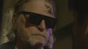 Een foto van een man met grijs haar en een baard. Hij heeft een zonnebril op en heeft tattoos van vlinders op zijn voorhoofd. De achtergrond is vrij donker.