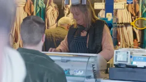 Een foto van een vrouw achter een toonbank. Het is de visboer. Er staan mensen in de rij. Op de achtergrond zie je allemaal bundels met touwen.