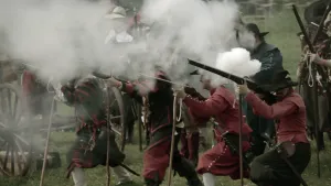 Een foto van een groep mannen die een oude veldslag naspelen. Je ziet oude geweren, die op stokken leunen, en daarmee schieten ze. Je ziet grote rookwolken van die geweren afkomen.