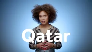 Een foto van een presentator die de vingertoppen van de linkerhand tegen de vingertoppen van de rechterhand houdt. Op de voorgrond staat de tekst "Qatar". Qatar is een land in het Midden-Oosten. De achtergrond is blauw van kleur.