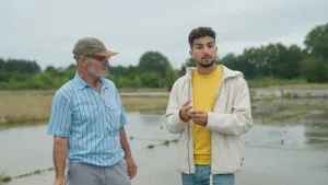 Een foto van de presentator met naast hem een oudere man van buitenlandse afkomst. Ze staan buiten en de presentator kijkt in de camera.