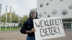 Een foto van een meisje dat voor een gebouw staat. Ze houdt een bord vast en kijkt in de camera. Op het bord staat de tekst "We kunnen beter!"