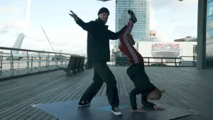 Een foto van Janouk die ondersteboven op haar handen staat. Haar benen worden in de lucht gehouden door een man. Ze staan buiten op een vierkante plaat, in Rotterdam.