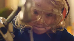 Een foto van een jongetje. Hij heet Dorus en heeft een veiligheidsbril en gehoorbescherming op. Hij slaat met een hamer op een spijker.