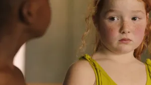 Een foto van het meisje Alisa. Ze heeft een badpakje aan en kijkt bang. Op de voorgrond zie je een jongetje deels in beeld.