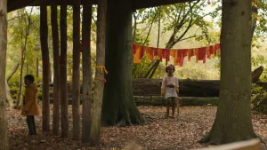 Een foto van een meisje van ongeveer tien jaar oud. Ze staat in een bos en heeft een houten zwaard bij zich. Aan een waslijn hangen allemaal gele en oranje lappen. Links in beeld staat een jongetje. 