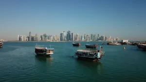 Een foto genomen in Qatar in het Midden-Oosten. Er zijn vijf motorboten te zien die over het water varen. Op de achtergrond is rechts een kleine aanlegstijger te zien en verder naar achteren zijn allemaal moderne, hoge gebouwen te zien, wolkenkrabbers.