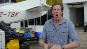 Een foto van bioloog Freek. Hij kijkt in de camera en is druk bezig om iets te vertellen. Hij beweegt daarbij met zijn armen. Achter hem zie je de staart van een vliegtuig. Ook zie je bagage op de grond staan.