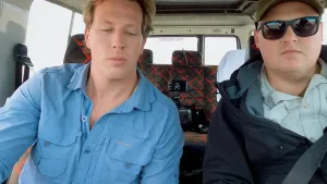 Een foto van bioloog Freek. Hij zit voor in een auto. Naast hem zit een man, dat is de bestuurder. Freek kijkt schuin naar beneden.