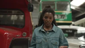 Een foto van presentator Sosha in een hal waar links een deel van een ouderwetse rode bus te zien is. Rechtsachter staat op een hoger deel een oude groene bus met twee voorruiten. Daaronder is een deel van een oude witte auto te zien.  