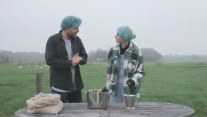 Een foto van Janouk en een man op een grasveld die een blauw netje op hun hoofd hebben. Voor hen staat een tafel met daarop een blauw bakje achter een witte zak met niet-zichtbare sojabonen, een pan met plastic staafmixer erin en een klein emmertje.