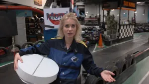 Een foto van Janouk in het midden van een kartbaan binnen met op de achtergrond karts en helmen. In haar rechterarm heeft ze een hele grote, ronde en witte pil met in het midden een gleuf, precies zoals een aspirine pil. Ze draagt een zwart met blauw pak.