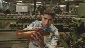 Een foto van presentator Nizar in de schoenenfabriek van Floris van Bommel. Hij toont een schoen aan de camera en wrijft daarbij over de zool. Op de achtergrond zie je allerlei machines en schoenen.