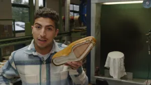 Een foto van Nizar die een deel van een schoen vastheeft in een fabriek. Hij houdt de onderkant van de schoen in de richting van de camera. Het lichtbruine leer van de bovenkant steekt onder uit en de onderkant met schoenzool zit nog niet op de schoen.